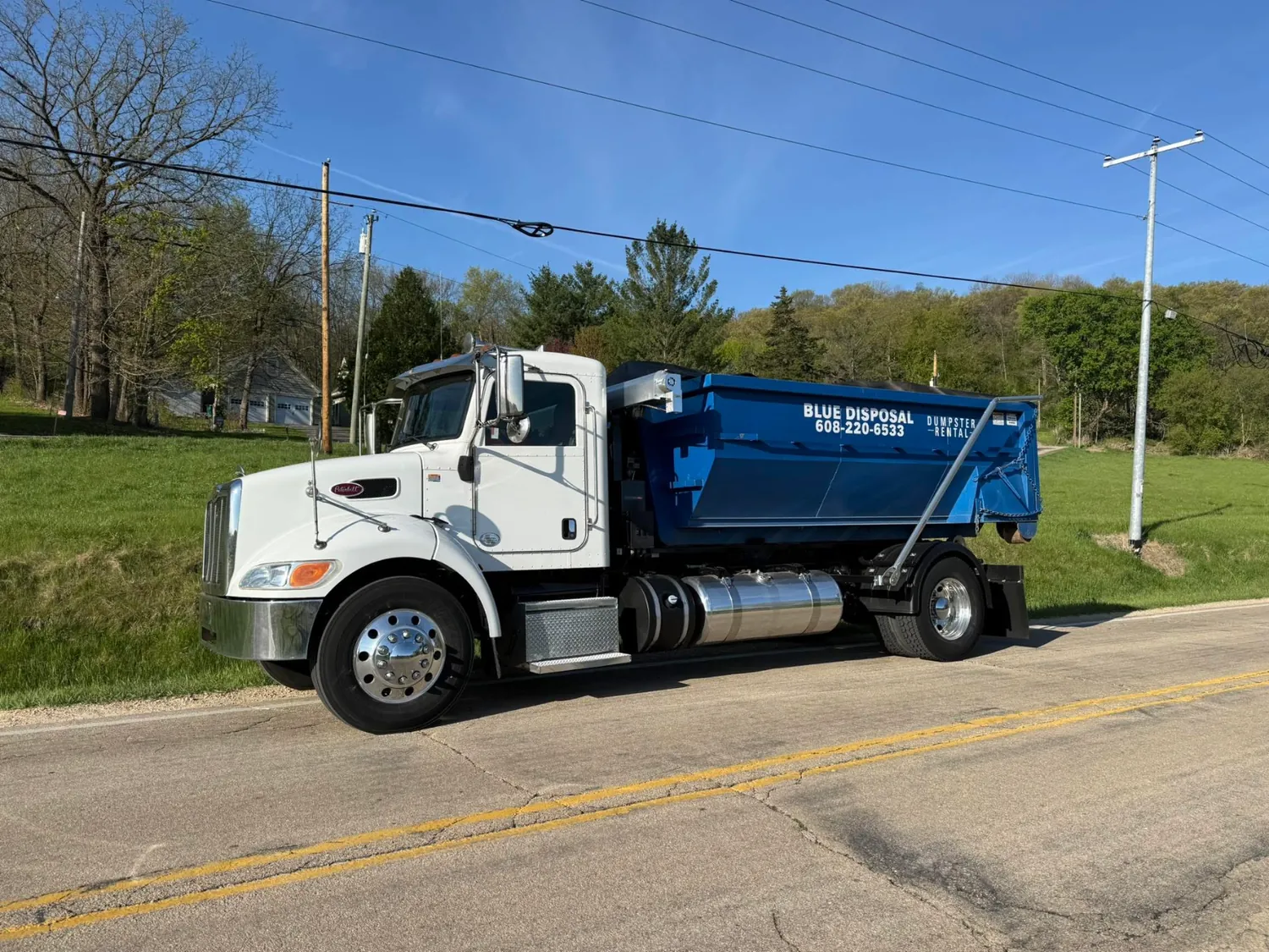 White roll-off truck carrying a blue Blue Disposal dumpster down a rural Wisconsin road